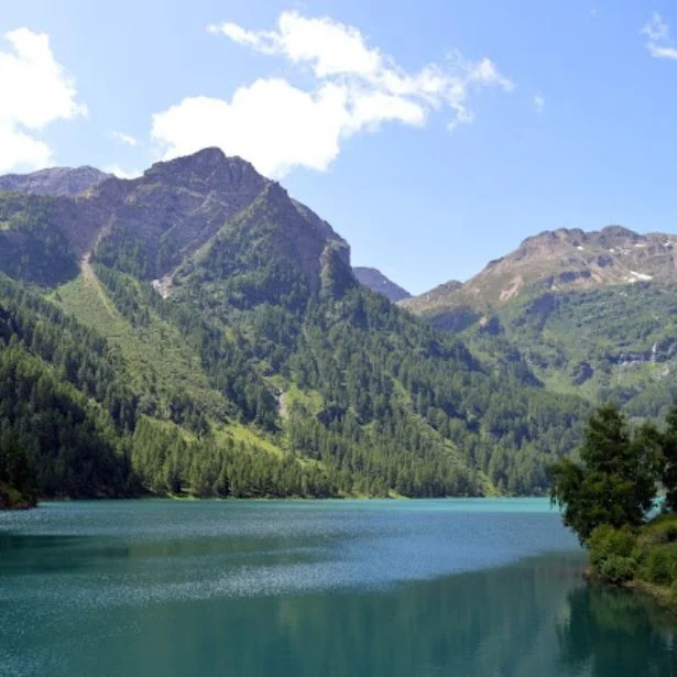 A calm blue-green lake reflects wooded mountains beneath a bright, partly cloudy sky.