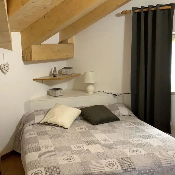 A compact bedroom with a double bed sits beneath exposed wooden beams, with shelves and dark curtains.