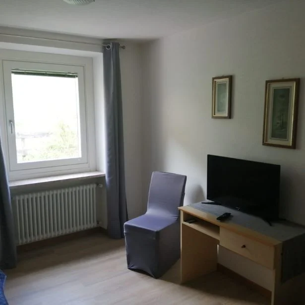 A simply furnished room contains a television on a cabinet, a covered chair and a window with gray curtains.