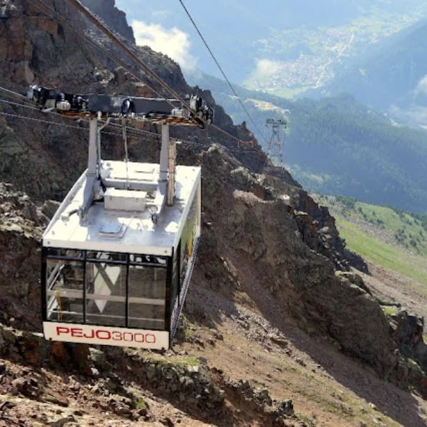 A cable car travels along a steep rocky mountainside above a green valley in bright daylight.